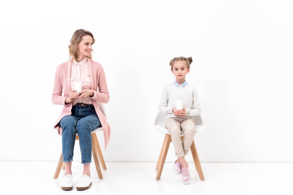 μητέρα και κόρη που κάθονται στις καρέκλες με ποτήρια γάλα σε λευκό - Φωτογραφία, εικόνα