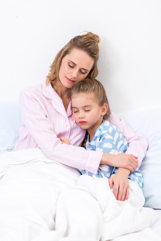 Mutter und Tochter sitzen mit geschlossenen Augen und umarmen sich zu Hause auf dem Bett - Foto, Bild