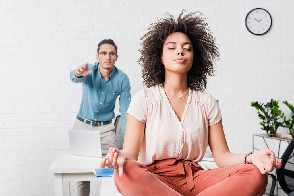 Молодая деловая женщина расслабляется и медитирует в офисе с коллегой-мужчиной позади
 - Фото, изображение