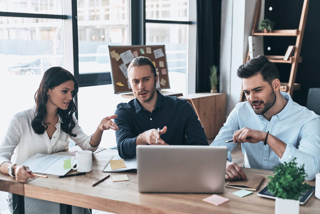 Успешная бизнес-команда. Группа молодых современных людей в элегантной повседневной одежде, указывая на ноутбук, сидя в креативном офисе
  - Фото, изображение