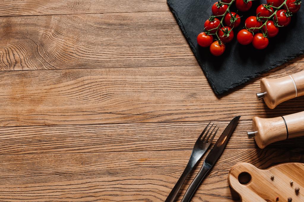 vista superior de tomates maduros frescos en pizarra, tenedor con cuchillo, condimentos en recipientes y granos de pimienta en mesa de madera
 - Foto, Imagen