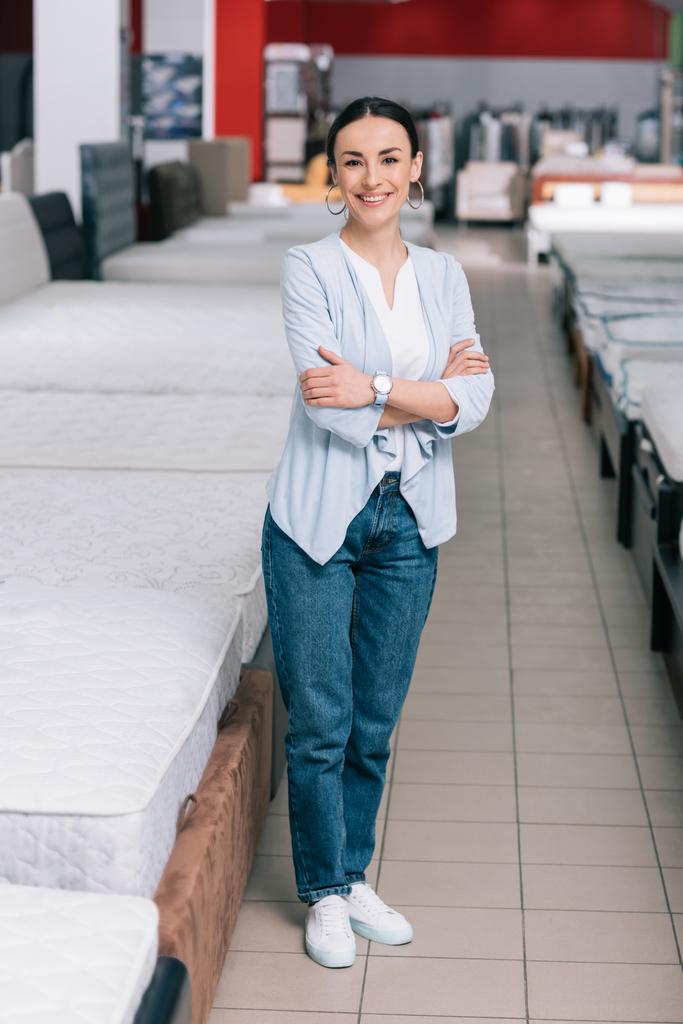 улыбающаяся клиентка со скрещенными руками, стоящая в мебельном магазине с расставленными матрасами
 - Фото, изображение
