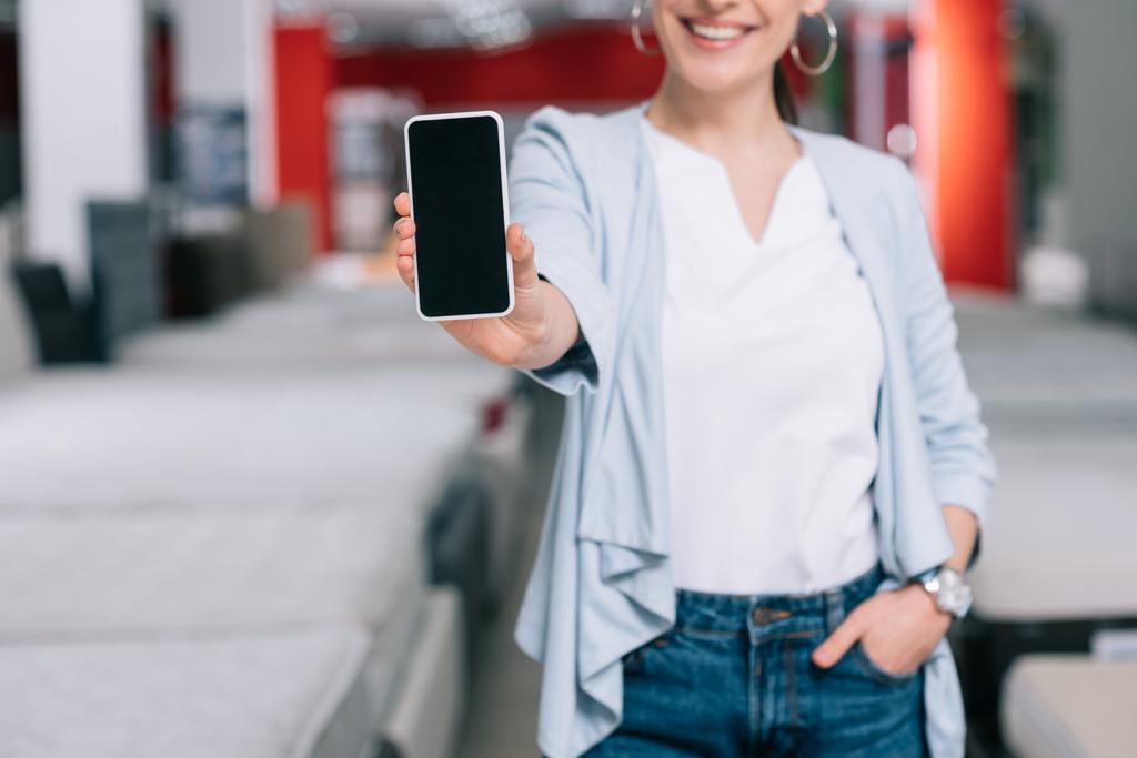Schnappschuss einer lächelnden Frau zeigt Smartphone mit leerem Bildschirm im Möbelhaus mit Matratzen - Foto, Bild