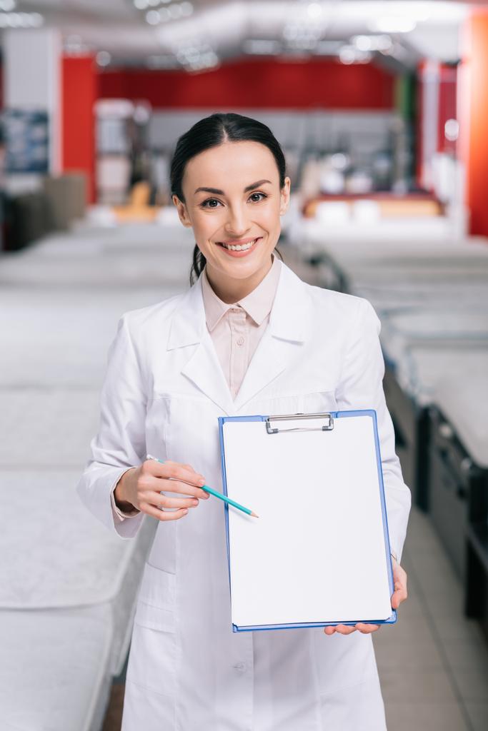 χαμογελώντας κατάστημα βοηθός σε λευκό παλτό δείχνει άδειο το σημειωματάριο στο χέρι στο κατάστημα επίπλων με τοποθετημένα στρώματα - Φωτογραφία, εικόνα