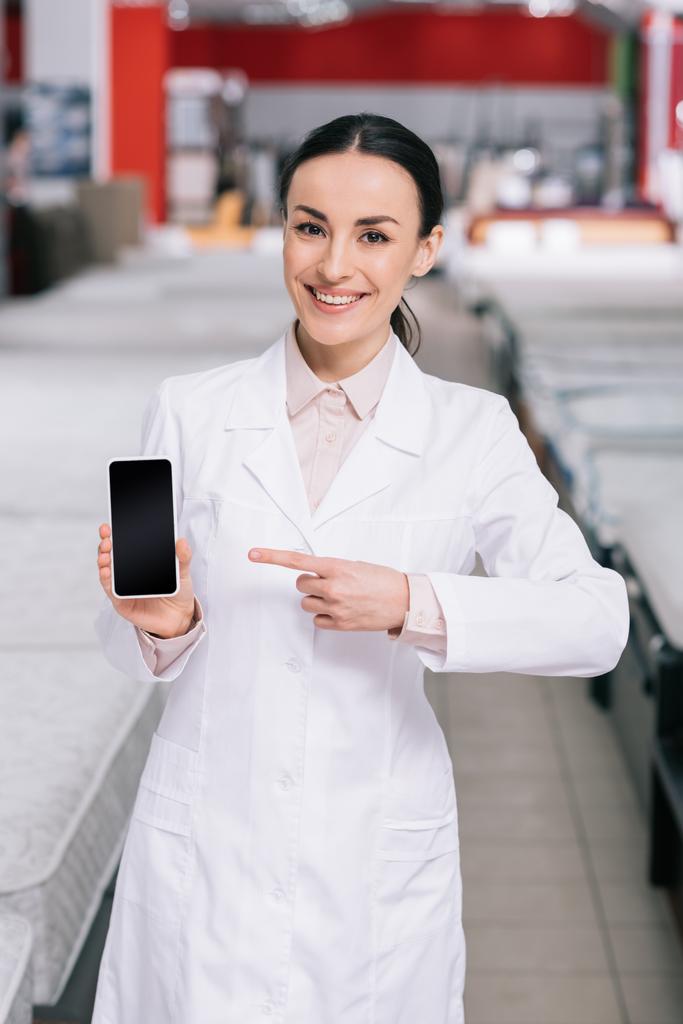 smiling shop assistant in white coat showing smartphone with blank screen in hand in furniture shop with mattresses - Photo, Image