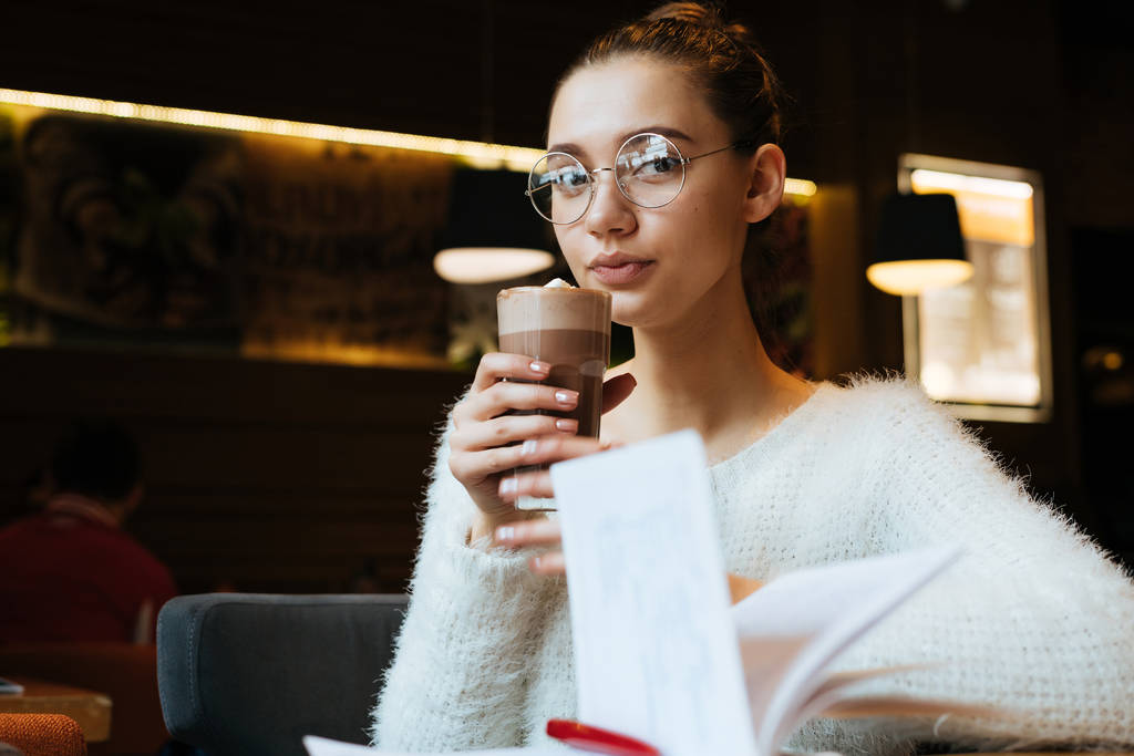 roztomilá mladá dívka student nosit brýle a bílé sako pití lahodné cappuccino v kavárně, po studiu - Fotografie, Obrázek