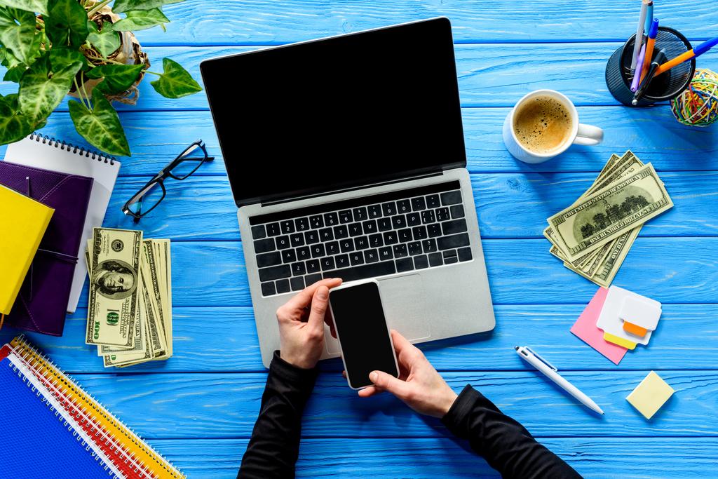 close-up view of person using smartphone by laptop on blue wooden table with stationery and money - Photo, Image