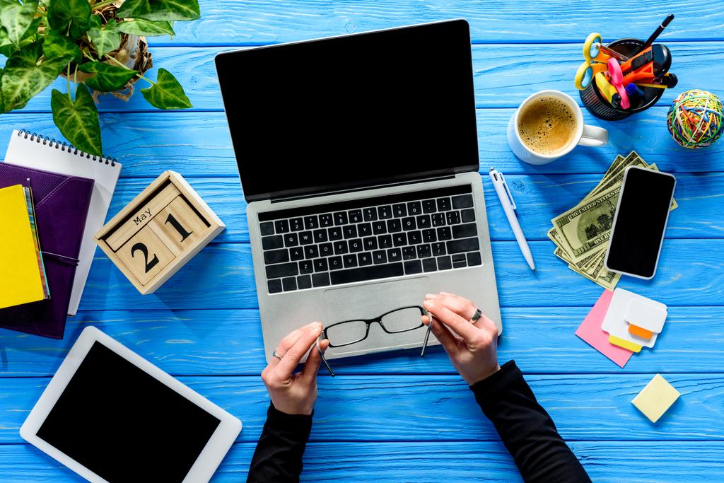 hands holding eyeglasses by laptop on blue wooden table with stationery and money - Photo, Image