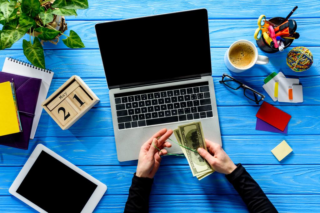 Geschäftsmann bindet Dollars mit Gummiband am Laptop auf blauem Holztisch mit Schreibwaren - Foto, Bild