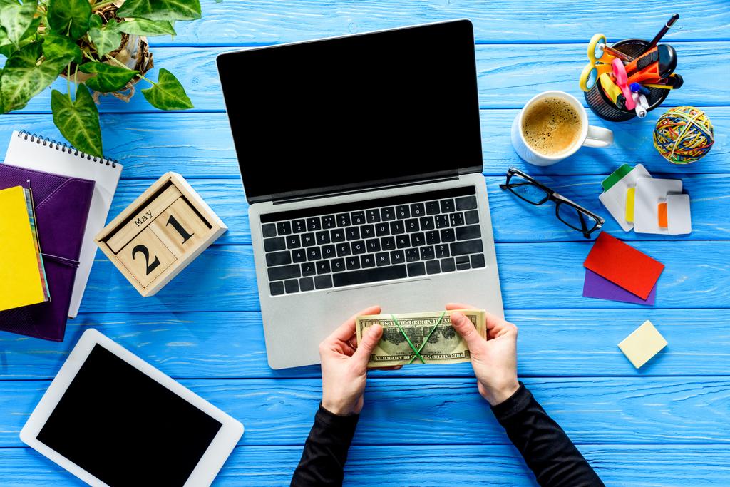 manos sosteniendo el dinero por ordenador portátil en la mesa de madera azul con papelería
 - Foto, imagen