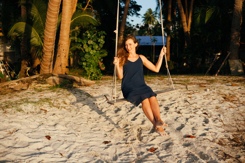 usmívající se krásná žena na houpačce mezi palmami při pohledu kamery na pláži - Fotografie, Obrázek