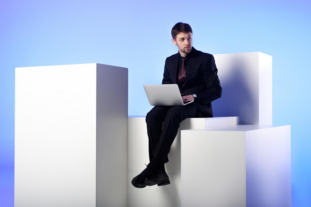 Geschäftsmann mit Laptop sitzt auf weißem Block isoliert auf weißem Block - Foto, Bild
