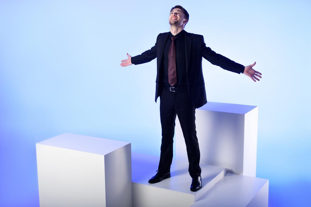 homem de negócios em terno preto com braços estendidos em pé no bloco branco isolado em azul
 - Foto, Imagem