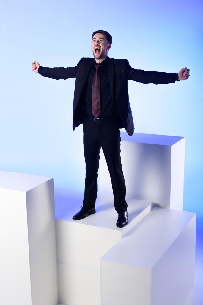 zakenman in zwart pak met uitgestrekte armen permanent op witte blok geïsoleerd op blauw - Foto, afbeelding