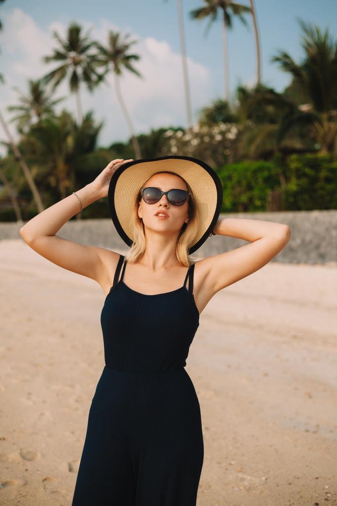 atractiva mujer posando en sombrero, gafas de sol y vestido en la playa de arena del océano
 - Foto, Imagen