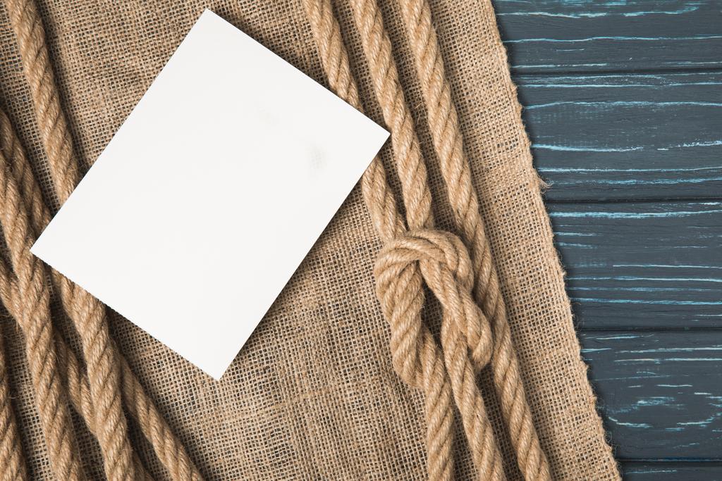 primer plano plano de papel blanco en blanco sobre tela de saco y cuerdas anudadas náuticas marrones sobre mesa de madera
 - Foto, Imagen