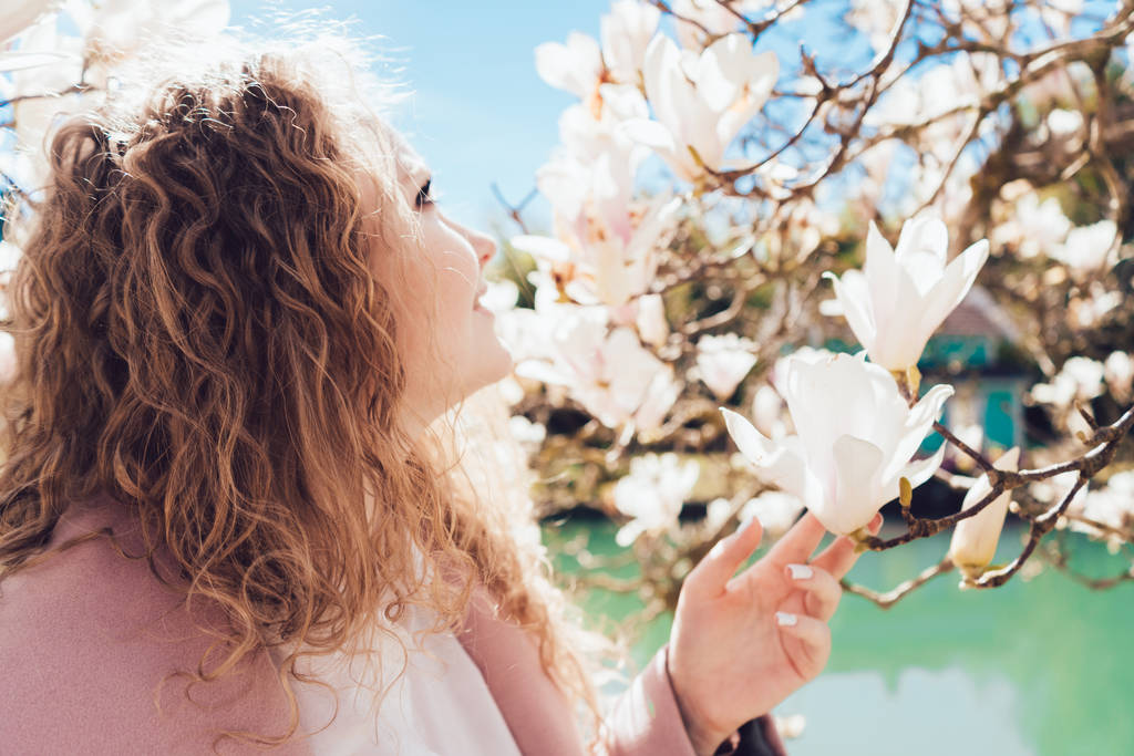 onnellinen kihara nuori nainen nauttia luonnosta, kukkivat magnolia puistossa taustalla lampi
 - Valokuva, kuva