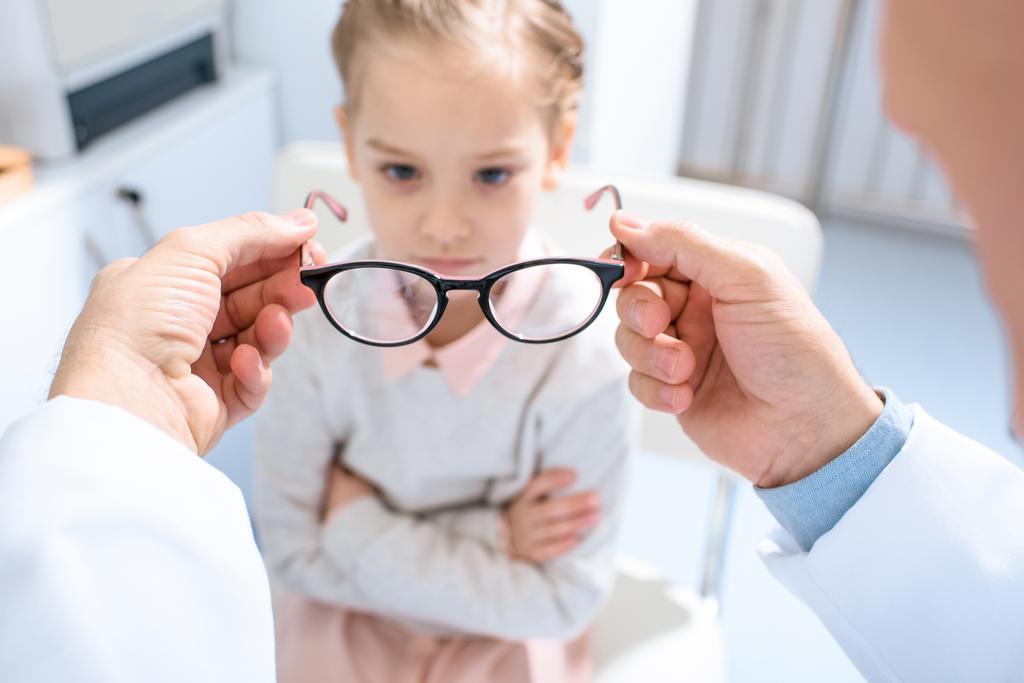 imagen recortada del oftalmólogo que propone gafas nuevas a un niño infeliz en la clínica
 - Foto, Imagen