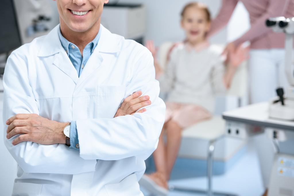 immagine ritagliata di oftalmologo sorridente in piedi con le braccia incrociate in sala di consultazione
 - Foto, immagini