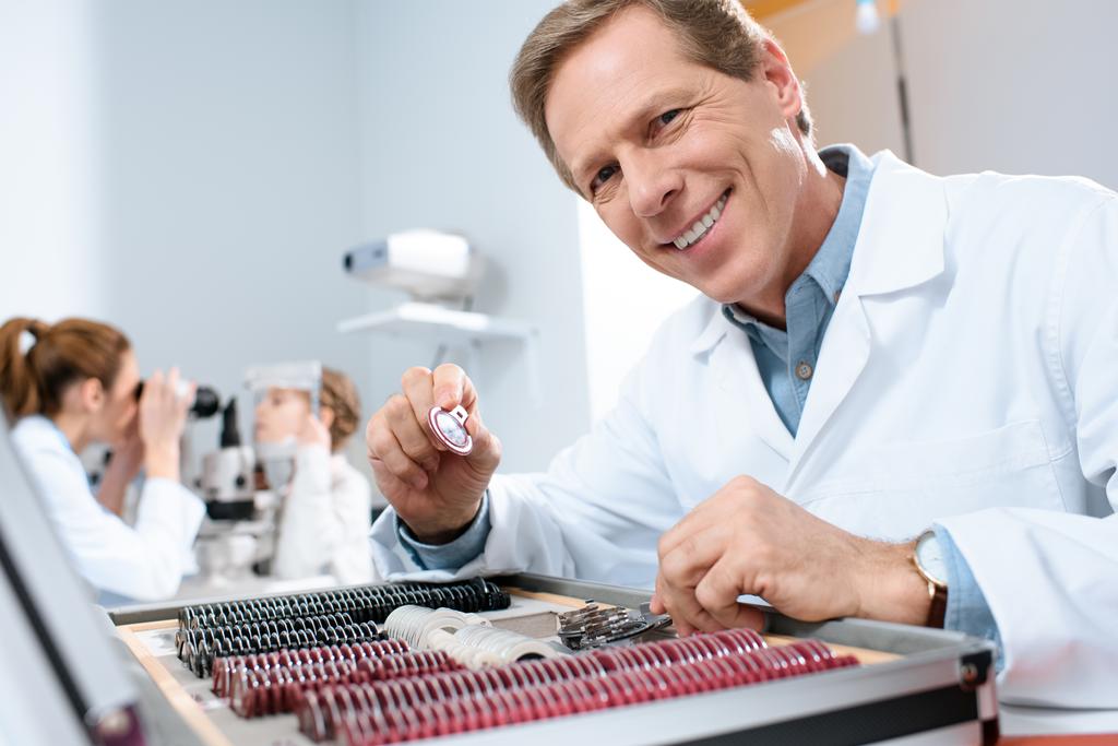 masculino sorrindo oftalmologista segurando lentes para quadro experimental enquanto colega examinando criança com teste de campo visual
 - Foto, Imagem