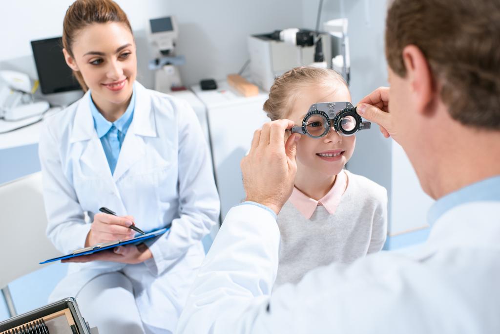 офтальмологи осматривают глаза ребенка с пробной рамкой и диагнозом
 - Фото, изображение