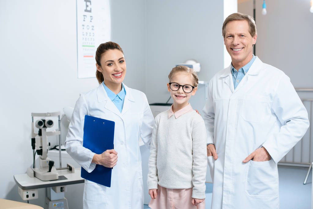 two smiling ophthalmologists and little child in glasses standing in clinic - Photo, Image