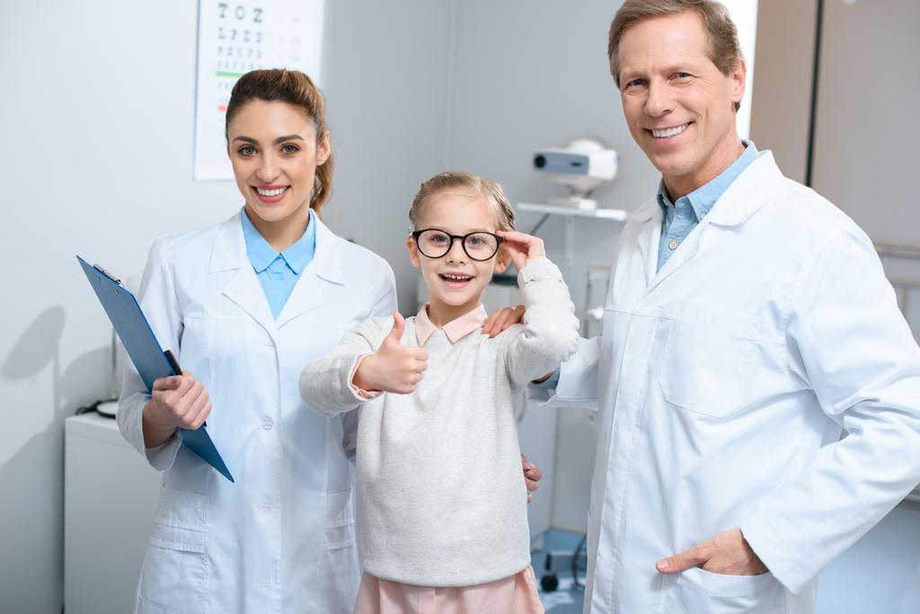 dois optometristas sorridentes e criança pequena em óculos mostrando polegar para cima
 - Foto, Imagem