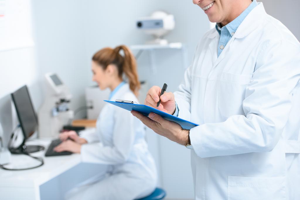 a férfi szemész írás diagnózis, miközben kollégája segítségével a számítógép a klinikán levágott megtekintése - Fotó, kép