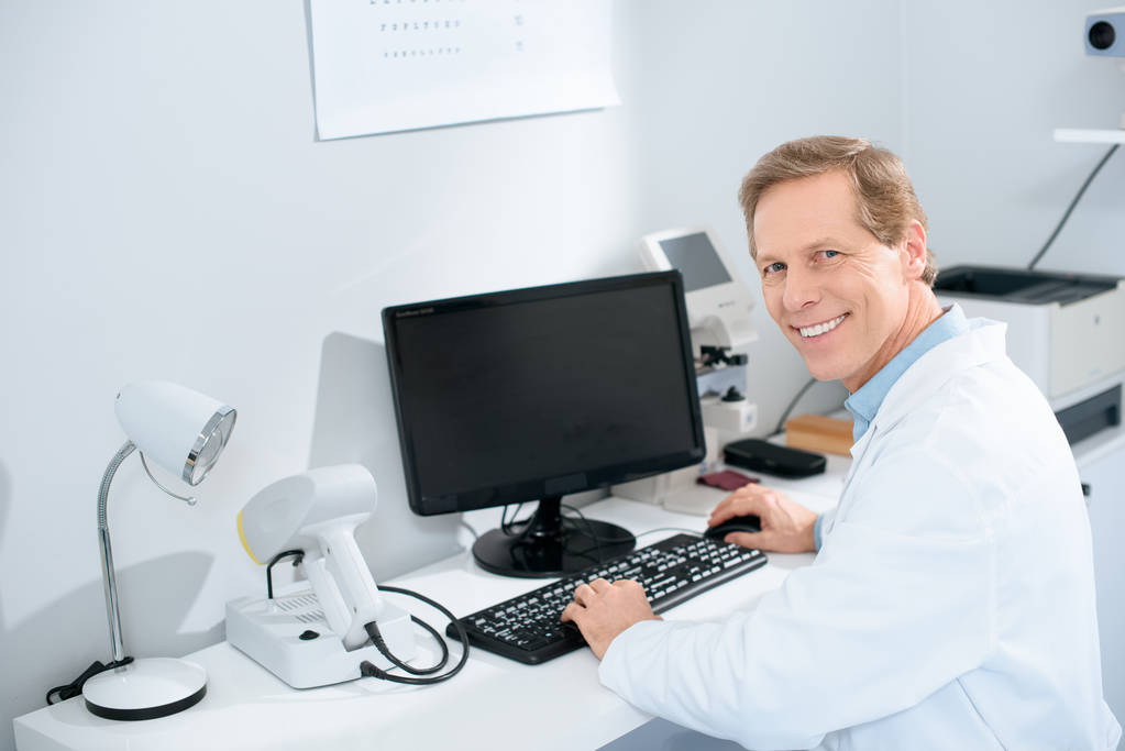 glimlachend mannelijke opticien werken met computer in kliniek - Foto, afbeelding
