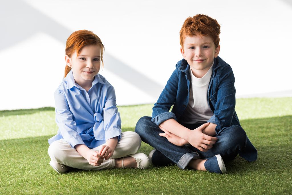 carino felice rossa bambini seduti insieme su erba e sorridente a macchina fotografica su grigio
  - Foto, immagini