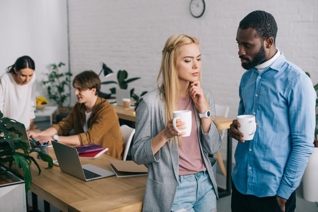 мультикультурні бізнес-партнери з чашками кави обговорюють і двоє співробітників працюють на ноутбуці позаду
 - Фото, зображення