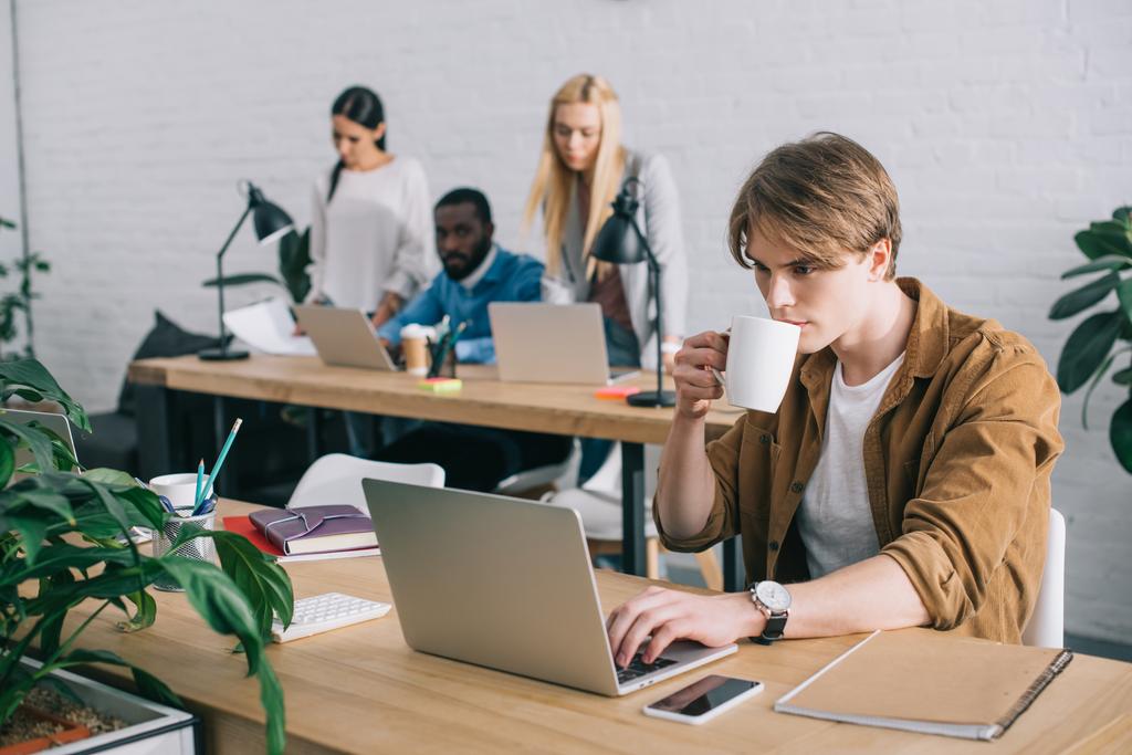 бизнесмен с кофейной кружкой с помощью ноутбука и коллеги, работающие в современном офисе
  - Фото, изображение