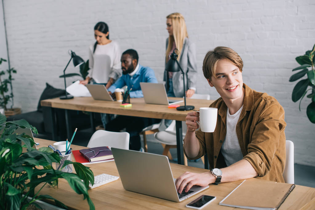 бізнесмен з чашкою кави за допомогою ноутбука та колег, що працюють позаду в сучасному офісі
  - Фото, зображення