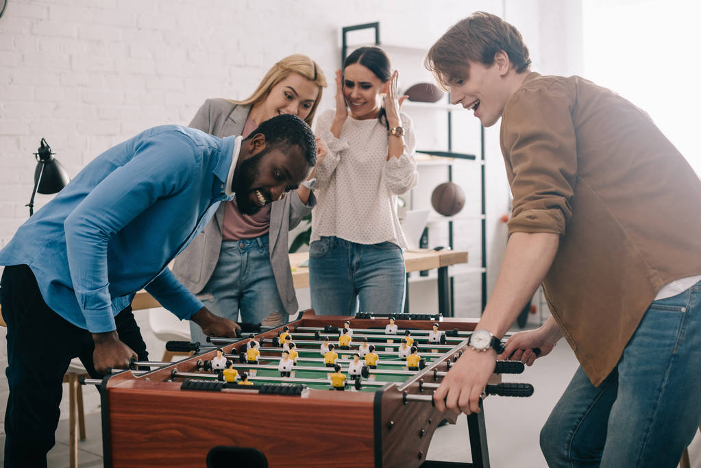 side view of happy multicultural businessmen playing table football in front of female colleagues  - Photo, Image