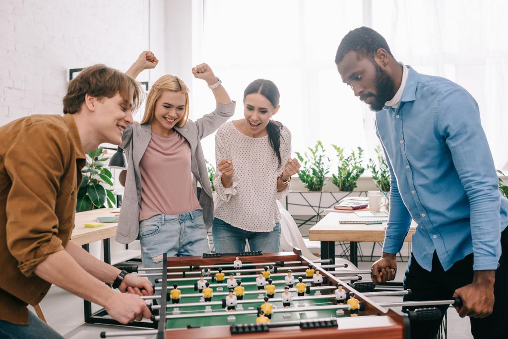 Zijaanzicht van gelukkig multiculturele zakenlieden tabel voetballen voor vieren en gebaren van vrouwelijke collega 's  - Foto, afbeelding