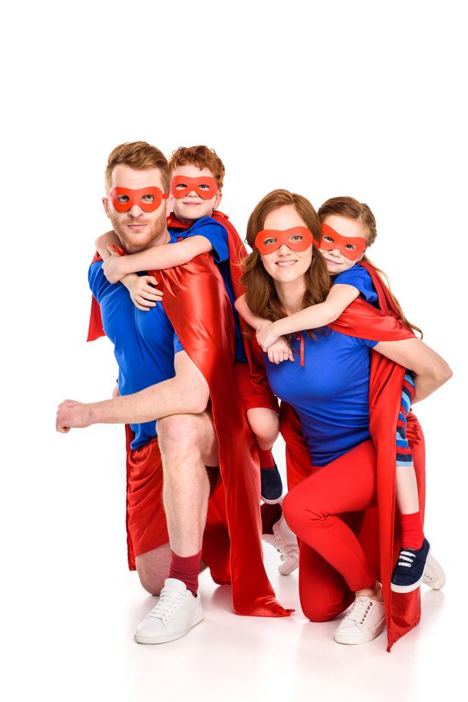 glückliche Supereltern huckepack entzückend lächelnde Kinder in Masken und Mänteln isoliert auf weiß - Foto, Bild
