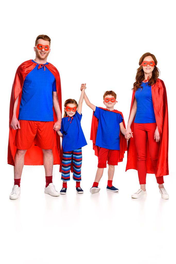 Ganzheitliche Ansicht einer glücklichen Superfamilie in Masken und Mänteln, die sich an den Händen hält und die Kamera isoliert auf Weiß betrachtet - Foto, Bild