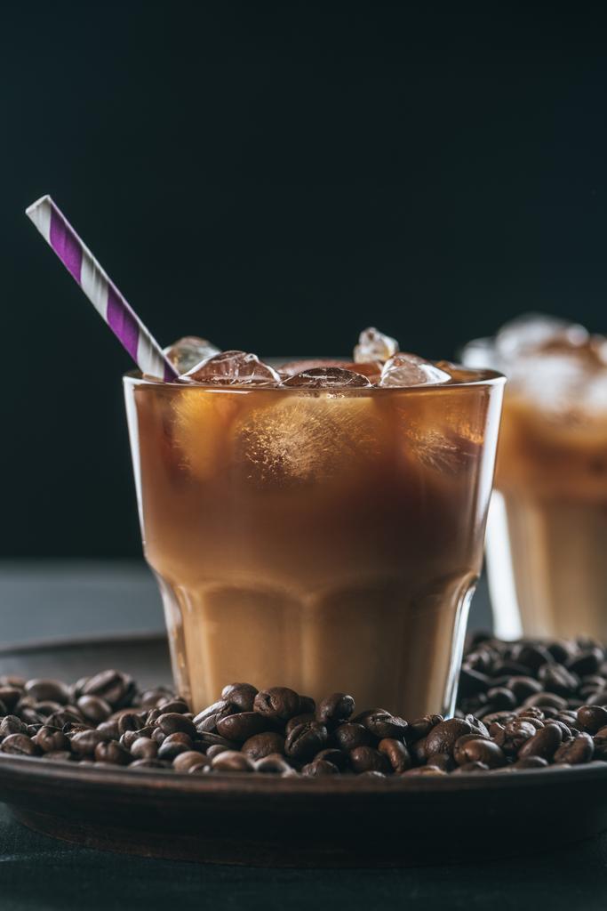 selektiver Fokus eines Glases kalten Eiskaffees mit Stroh auf dem Teller mit gerösteten Kaffeebohnen auf der Tischplatte vor dunklem Hintergrund - Foto, Bild