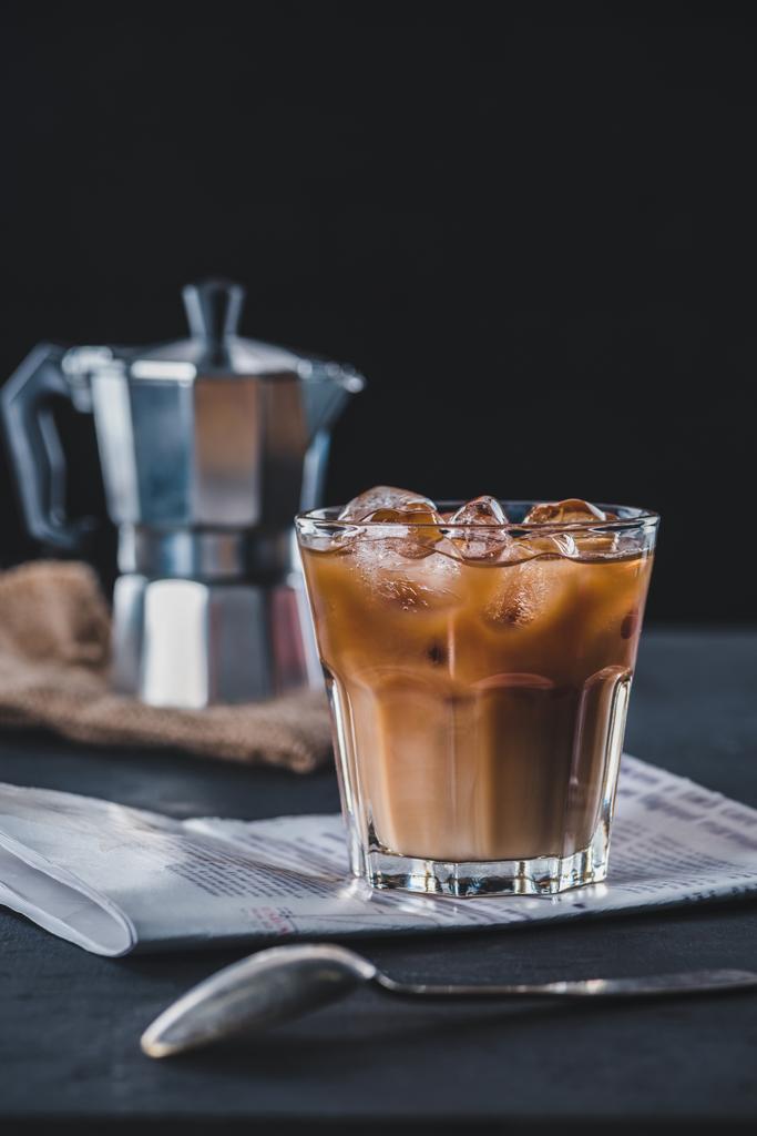 επιλεκτική εστίαση ποτήρι κρύο παγωμένο καφέ, εφημερίδα και καφετιέρα εσπρέσο σε επιτραπέζια σε σκούρο φόντο - Φωτογραφία, εικόνα