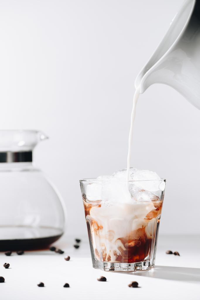 Nahaufnahme des Einfüllens von Milch in Glas mit kalt gebrühtem Kaffee, Kaffeemaschine und gerösteten Kaffeebohnen auf grauem Hintergrund - Foto, Bild