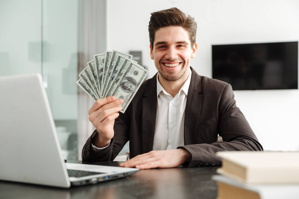 Χαρούμενος νεαρός επιχειρηματίας δείχνει χρήματα που χρησιμοποιούν φορητό υπολογιστή. - Φωτογραφία, εικόνα
