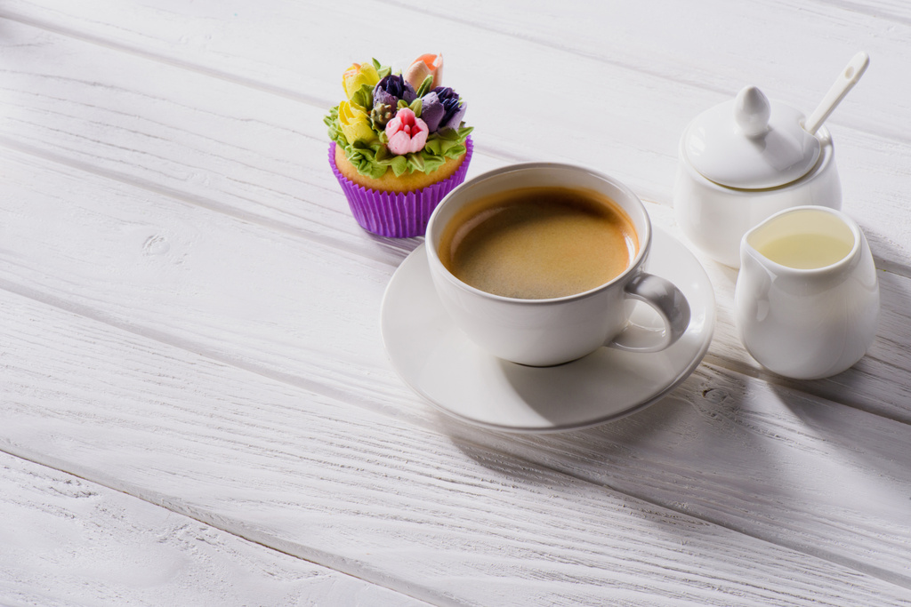 Nahaufnahme von Tasse Kaffee, süßem Muffin und Sahne auf weißer Holzplatte - Foto, Bild