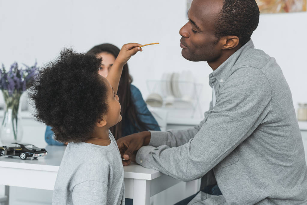 african american son feeding father with crispy breadsticks at home - Photo, Image