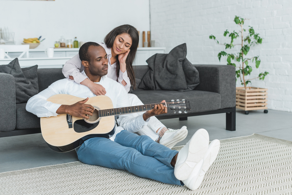 african american boyfriend sitting on floor and playing guitar for girlfriend at home - Photo, Image