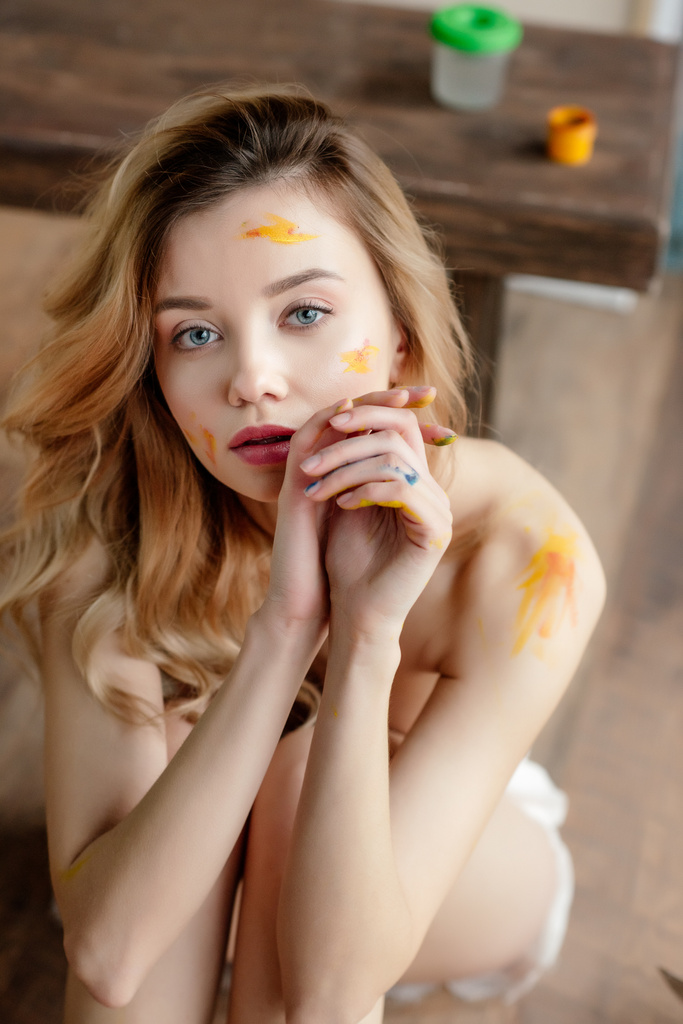 high angle view of beautiful sensual naked girl with paint on face and body sitting and looking at camera - Photo, Image