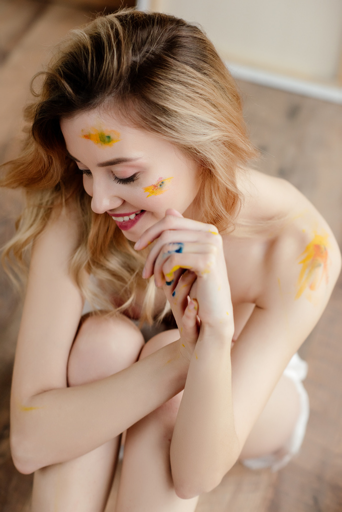 hoge hoekmening van mooie sensuele naakt meisje met paint op gezicht en lichaam zitten op verdieping van kunststudio glimlachen - Foto, afbeelding