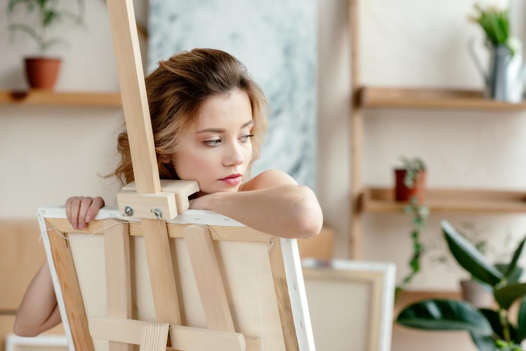 kaunis aistillinen nuori nainen nojaa maalaustelineeseen ja katselee pois taidestudiossa
 - Valokuva, kuva