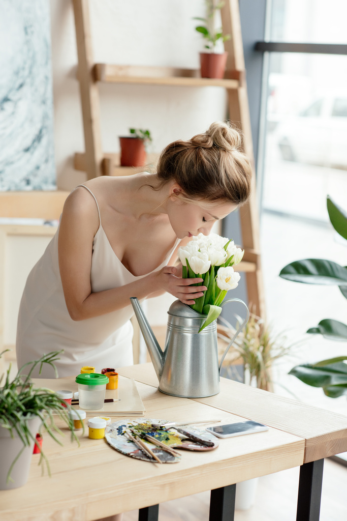 beautiful young woman sniffing flowers in watering can in art studio - Photo, Image