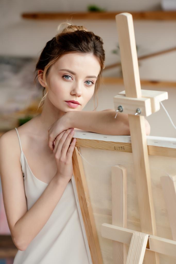 schöne junge Frau lehnt an Bild auf Staffelei und blickt in Kamera im Kunstatelier - Foto, Bild