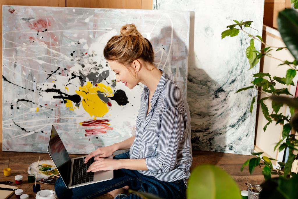 vista lateral de la joven pintora sonriente usando el portátil en el estudio de arte
 - Foto, Imagen